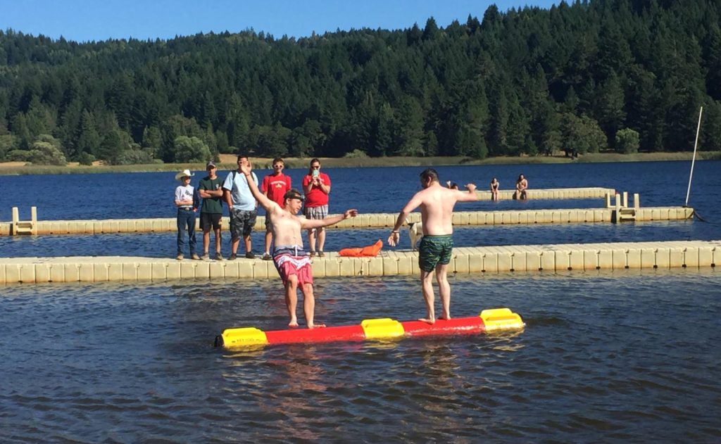 Two scouts competing in Campwide Games in the Log Rolling event at Waterfront, Wente Scout Reservation with other on the docks in the background.